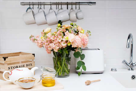 wedding bouquet of pink and orange lisianthus, antirrhinum and eucalyptus in glass vase on kitchen table. Black tea in a white cup, honey jar and spoon for honey, lemon closeup on a white kitchenの写真素材