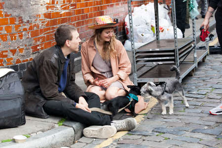 London, UK - 17 July 2019, Columbia Road Flower Market. A girl in a pink cloak and gray short silk overalls and a straw hat sits on the sidewalk with her friend, looks at the terrier puppy on a leash.のeditorial素材