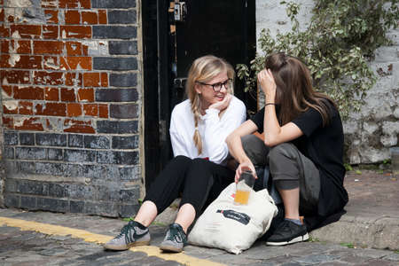 London, UK - 17 July 2019, Columbia Road Flower Market. Two girls with beer in their hands are sitting on the sidewalk, laughing and chatting.のeditorial素材