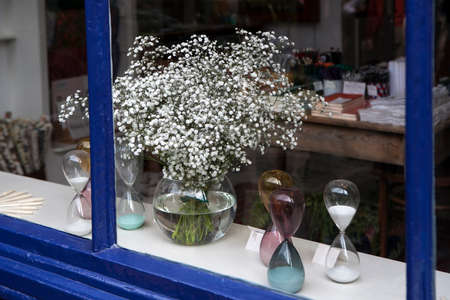 London, UK - 17 July 2019, Bouquet of gypsophila in a round glass vase in a store window with an hourglassのeditorial素材