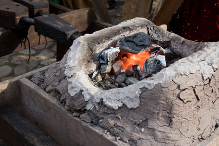 A blacksmith sets up the brazier with embers with large coal tongs. Embers glow in a iron forge....