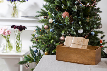 Wrapped in craft beige paper, the tied gifts are folded into a wooden box. They lie on the table against the background of a decorated Christmas tree. Copy spaceの写真素材
