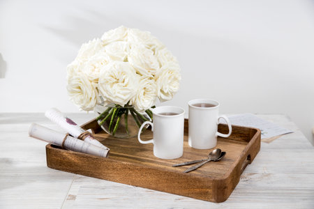 A bouquet of white roses in a round glass vase on a tray with two cups of tea and spoons in the interior. The letters with pictures are rolled up and tied with twine .. Copy spaceの写真素材