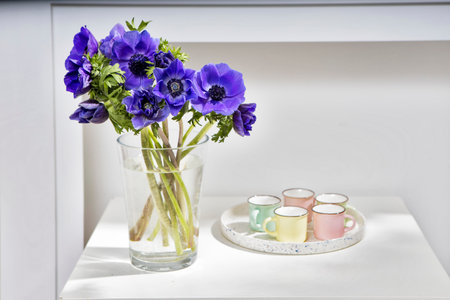 bouquet of the blue anemones in a glass vase on a white table near the pale gray wall. Tray with five small cup for americano. Vertical frame. Copy space.の写真素材