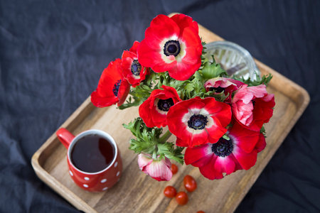 A bouquet of red anemones in a glass vase on a wooden tray on the bed. White polka dot cup with tea....