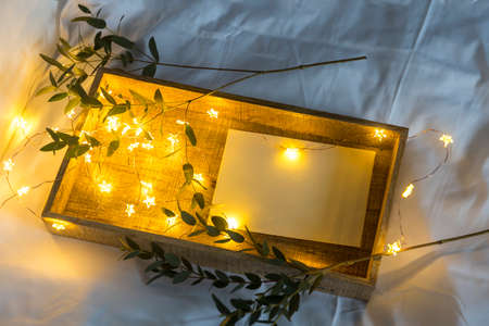 A eucalyptus branch lies on a white sheet beside a tray with a letter in an envelope. The garland is...