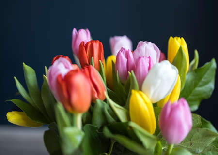 A bouquet of multi-colored tulips in a transparent vase on the tall chair with step against a dark...