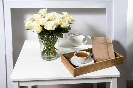 A bouquet of white roses in a vintage glass vase, a wooden tray with a cup of tea, a faience...