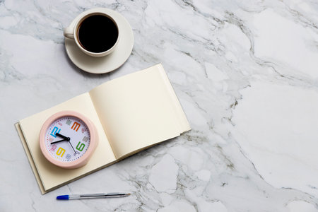 Round pink clock, square notebook, and pen on marble table surface. Cup of coffee. Copy spaceの写真素材