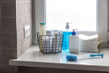 A used bottle of body lotion, blue liquid soap stands on a windowsill in a bathroom with beige tiles. There are small white terry hand towels in a black mesh metal basket. Washing brush.の写真素材