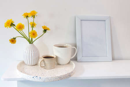 A bouquet of dandelions in a white fluted vase and two cups of different sizes with coffee on a white table and frame for picture. Breakfast. Copy spaceの写真素材