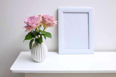 One pale purple rose in a white fluted vase and picture frame with space for text on the table as...