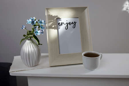 Flower hackelia velutina in a white fluted vase in the style of the seventies, a photo frame with the words Enjoy, and a cup of coffee on the dresser. Scandinavian styleの写真素材