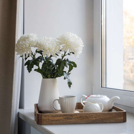 Three white chrysanthemums in a seventies-style fluted vase. Wooden tray with a kettle, milk jug and a cup of tea on the windowsill. Morning breakfast.の写真素材
