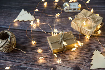 Glowing garland star, two gray boxes with gifts tied with twine, plywood Christmas decorations carved from wood are on a wooden table surfaceの写真素材