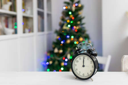A black alarm clock on a white table shows twelve o'clock. In the background there is a Christmas...