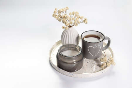 A vase with white dry flowers, a gray enamel cup with a drawn heart with tea, a burning candle stand...