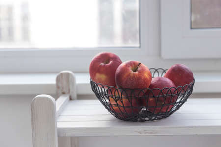 Red apples in a black mesh basket are on a white wooden shelf. Space for text. Copy placeの写真素材