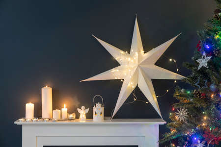 Home decoration before Christmas. Decorated Christmas tree with garland lights, rocking horse, gifts wrapped in elegant paper, artificial fireplace with a large white paper star and candles.の写真素材