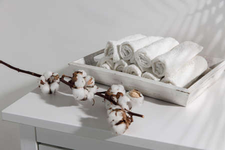 A wooden tray with rolled face towels and a branch of cotton flowers on a chest of drawers in the bathroom. The shadow of a palm leaf. Bathroom decoration for Easter. Place for textの写真素材