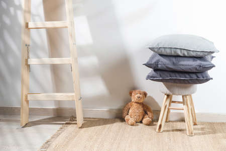 The teddy bear is leaning against the wall next to a small children's stool with three gray pillows on it. The stepladder is leaning against the wall. Copy spaceの写真素材
