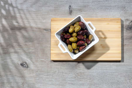 Deep square porcelain bowl with green and dark olives on a wooden board with shadow from a palm tree on a blue backgroundの写真素材