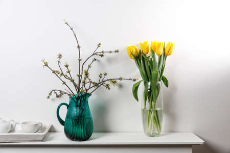 Bouquet of yellow tulip in the glass vase and American maple branches in green glass jar on white tableの写真素材
