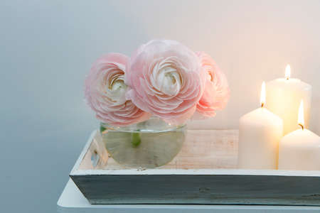 Three pale pink ranunculus in a transparent round vase and candles on the white windowsill....