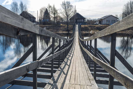 Verkhovye, Olonetsky district, Karelia, Russia, - April 28, 2022, Narrow wooden pedestrian suspension bridge across the river in russian village near Olonets. Sunny summer evening.の写真素材