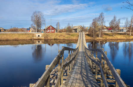 Verkhovye, Olonetsky district, Karelia, Russia, - April 28, 2022, Narrow wooden pedestrian suspension bridge across the river in russian village near Olonets. Sunny summer evening.のeditorial素材