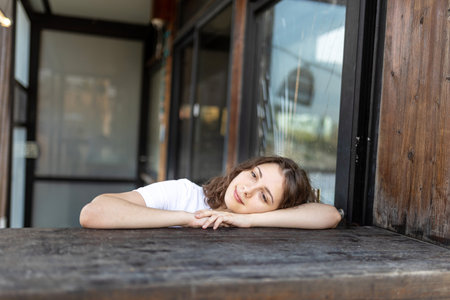 A smiling brunette girl in a white T-shirt, blue jeans sits at a wooden counter in a cafe, resting her chin in her hands. Vertical frame.の写真素材