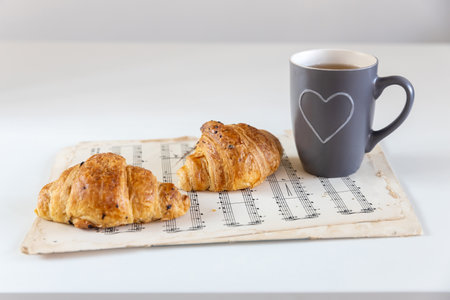 Two croissants with chocolate chips on a white sheet of paper on the table, a gray cup with a white heart with tea. Place for textの写真素材