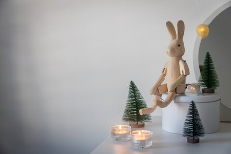 A hinged wooden hare, a small artificial green Christmas tree and lighted candles in candlesticks on a white chest of drawers. Space for text. Christmas card. vertical frameの写真素材