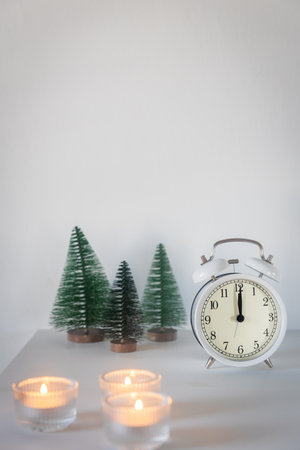 A white alarm clock, a twelve pointer, a small artificial green Christmas tree and lighted candles in candlesticks on a white chest of drawers. Copy space. Christmas card.の写真素材