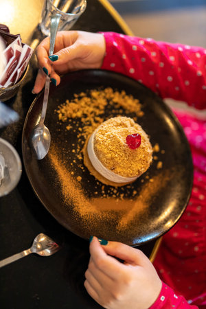 Cake with custard and cherry on top on a black plate, sprinkled with crumbs. Girl's hands with green...