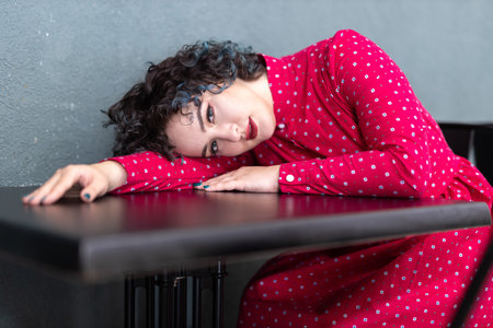Portrait of a smiling brunette girl of twenty-five years old dressed in a vintage red dress with a pattern with a small purse bag sits at a table in a cafe with a green wall.の写真素材