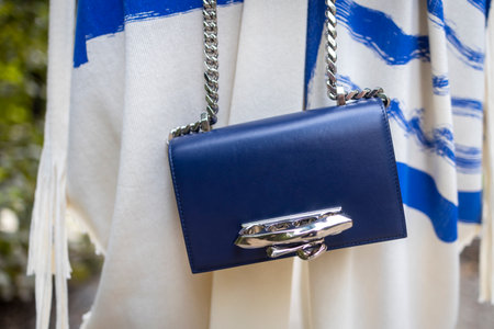 A small blue baby bag made of genuine leather on a silver chain is held by men's hands. Against the background of a white and blue jumpsuit in a geometric patternの写真素材