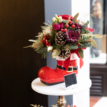 Christmas bouquet of gerbera, red tulip, white rose, pine cone, golden asparagus in a red Santa's boot instead of a vase on a red table in front of the entrance to a flower shopの写真素材