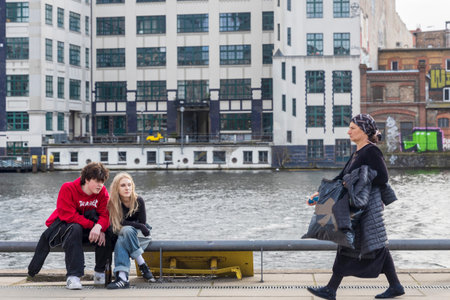 Berlin, Germany - 02 April 2024, A young guy and a blonde girl in a red hoodie are sitting on the embankment on the railing near the river.のeditorial素材