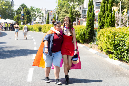 Haifa, Israel June 21, 2024, Pride Parade. Two girls wrapped in rainbow flags walk hugging each other along the road. One is wearing a beautiful short dress. The other is wearing jeans.のeditorial素材