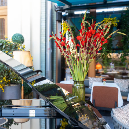 A bouquet of gladiolus in a clear glass vase between tables on the veranda, reflected in the lid of a grand piano. A yellow vintage table lamp glows. Cozy atmosphere in anticipation of guests.の写真素材
