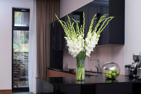 A white bouquet of gladioluses on the kitchen table in a tall glass vase with a bowl of green apples.の写真素材