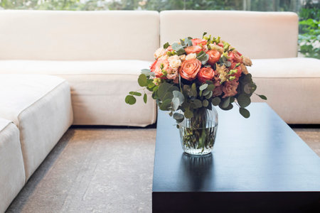 A bouquet of orange roses, pink and green lisianthus, and eucalyptus branches stands on a coffee table near a white sofa facing the window. Minimalism. Interior decoration.の写真素材
