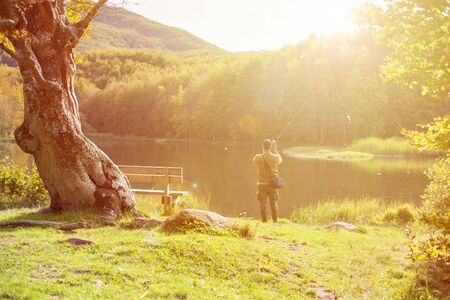 fisherman while casting line to fish in a mountain lake surrounded by greenery during sunsetの写真素材