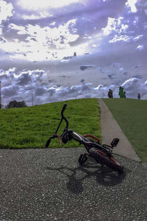 An abandoned bicycle lies on the path at the bottom of a grassy slope in the kids play area at the park on a warm sunny bright dayの写真素材