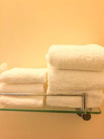 Fresh towels folded on a glass shelf under a warm light against a pale backgroundの写真素材