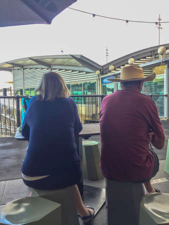 Sunday Dec 16th 2018, Auckland New Zealand - A man wearing a hat and a woman sit in silent companionship on stools with their backs turned at an outdoor cafe.のeditorial素材