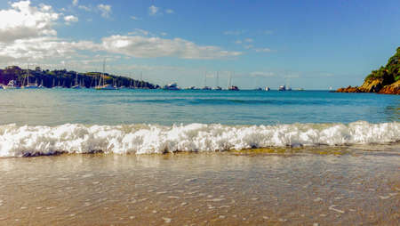 Sail boats with their mast sticking up against the blue sky with a foamy wave crashes to the shore in the foregroundの写真素材