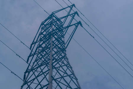 Upward view of an electrical pylon tower with cables running diagonally under a dull blue skyの写真素材