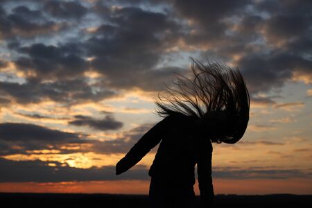 The silhouette of a young girl feels happy and shakes her head. The moment of sunset. Long hair loose. Shadow of a girl on a sunset background.の写真素材